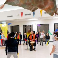 A large circle of children dancing and throwing colorful scarves while someone drums in the center.