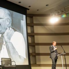 A stage is set up with two chairs on a rug with microphones for a discussion with a screen showing Uri Herscher and Howard Friedman.