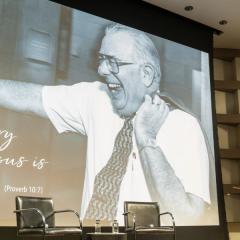 A stage is set up with two chairs on a rug with microphones for a discussion with a screen showing Uri Herscher and Howard Friedman.