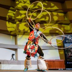 Two Native Hoop performers dance with multiple, interlocking hoops dressed in traditional clothing on a stage.