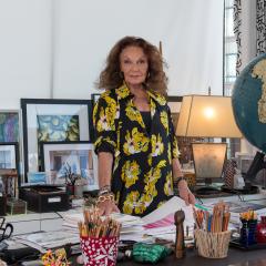 A woman with brown hair wearing a black and yellow dress stands behind a desk