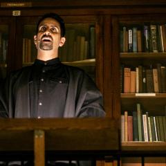 Still image of a performance featuring a man speaking in front of a tall bookcases filled with old, hardbound books.