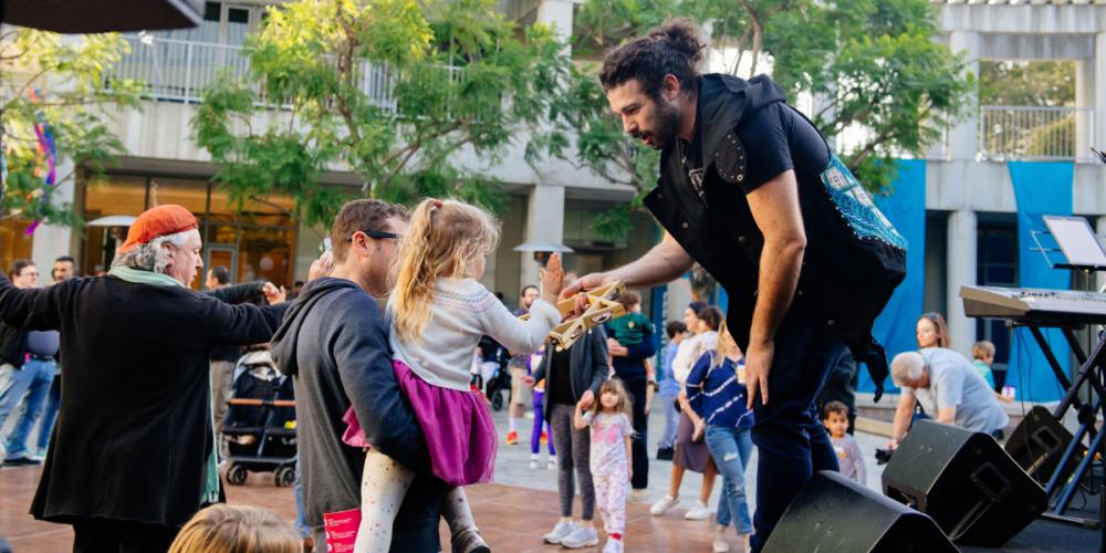 During a crowded celebration, a young child is held up by their adult to touch a tambourine in the shape of the Star of David that’s being held by a musician on a stage.