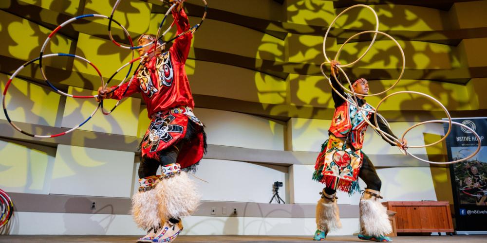 Two Native Hoop performers dance with multiple, interlocking hoops dressed in traditional clothing on a stage.